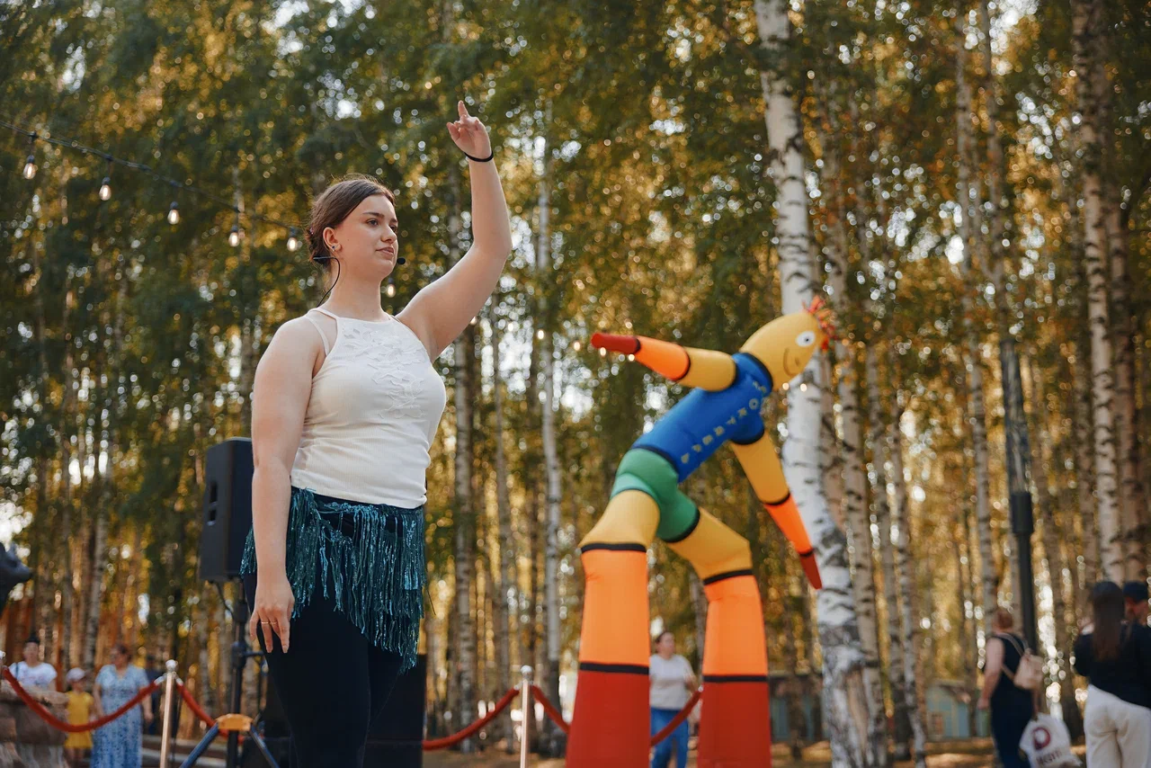 На фото женщина стоит в лесу на площадке и поднимает руку, рядом видны большие цветные фигуры и декорации, вокруг деревья и люди.