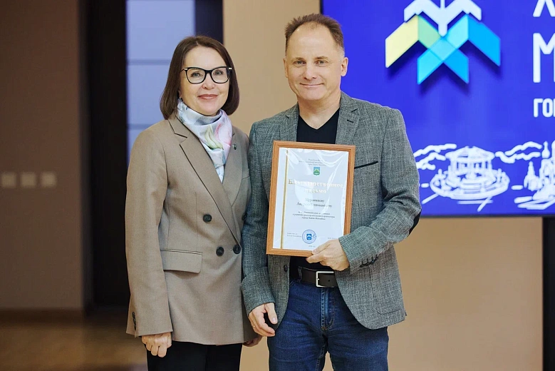 На фото двое людей стоят рядом. Мужчина держит сертификат в рамке, женщина рядом. На фоне экран с надписью «Ханты‑Мансийск».