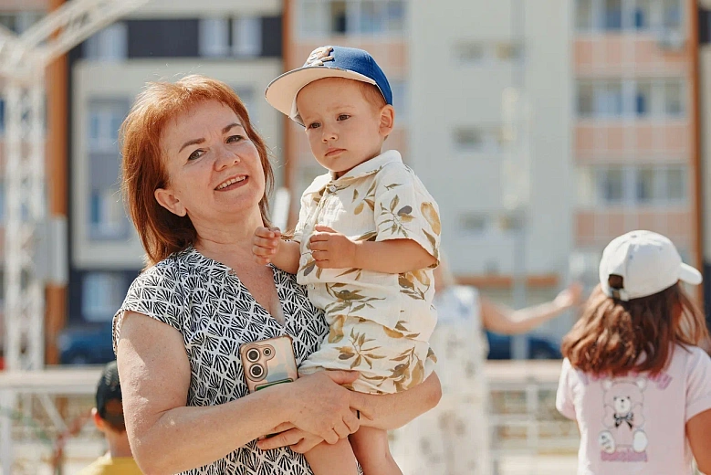 Женщина держит на руках маленького ребёнка, улыбается. На заднем плане дети стоят возле здания.